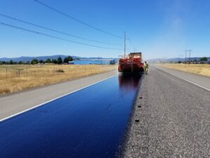 Asphalt truck performing road maintenance in rural Oregon under a clear sky.