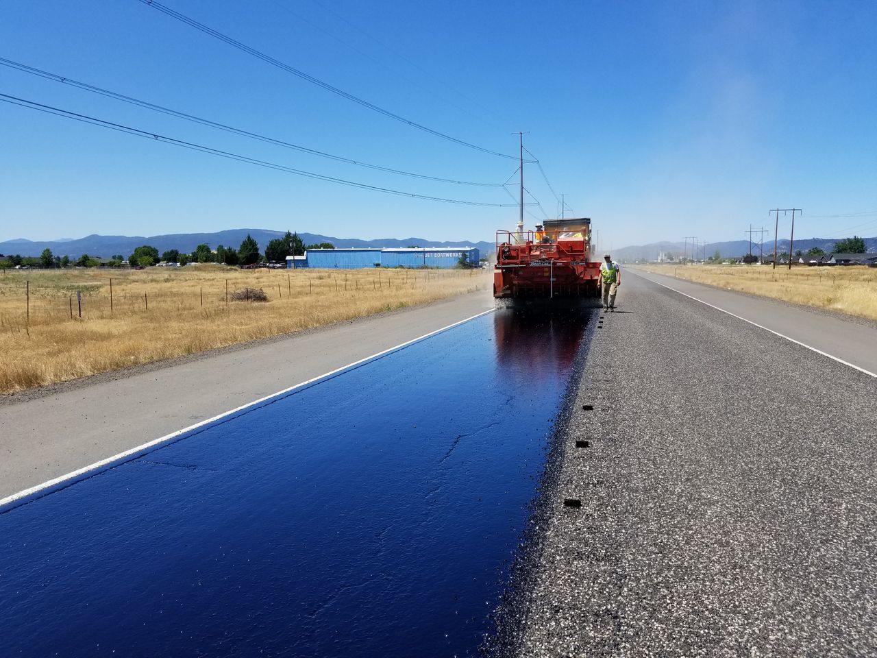 Asphalt truck performing road maintenance in rural Oregon under a clear sky.